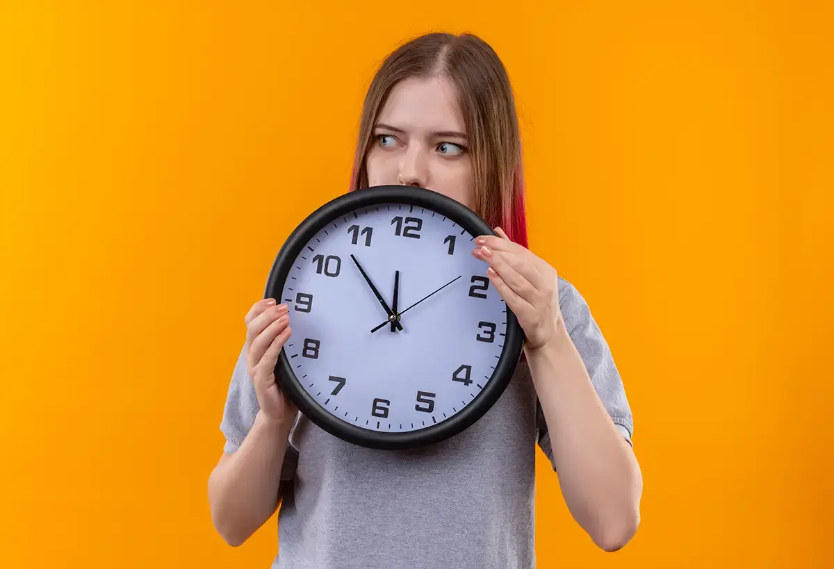 imagem com fundo sólido laranja e uma mulher de camiseta cinza segurando um grande relógio